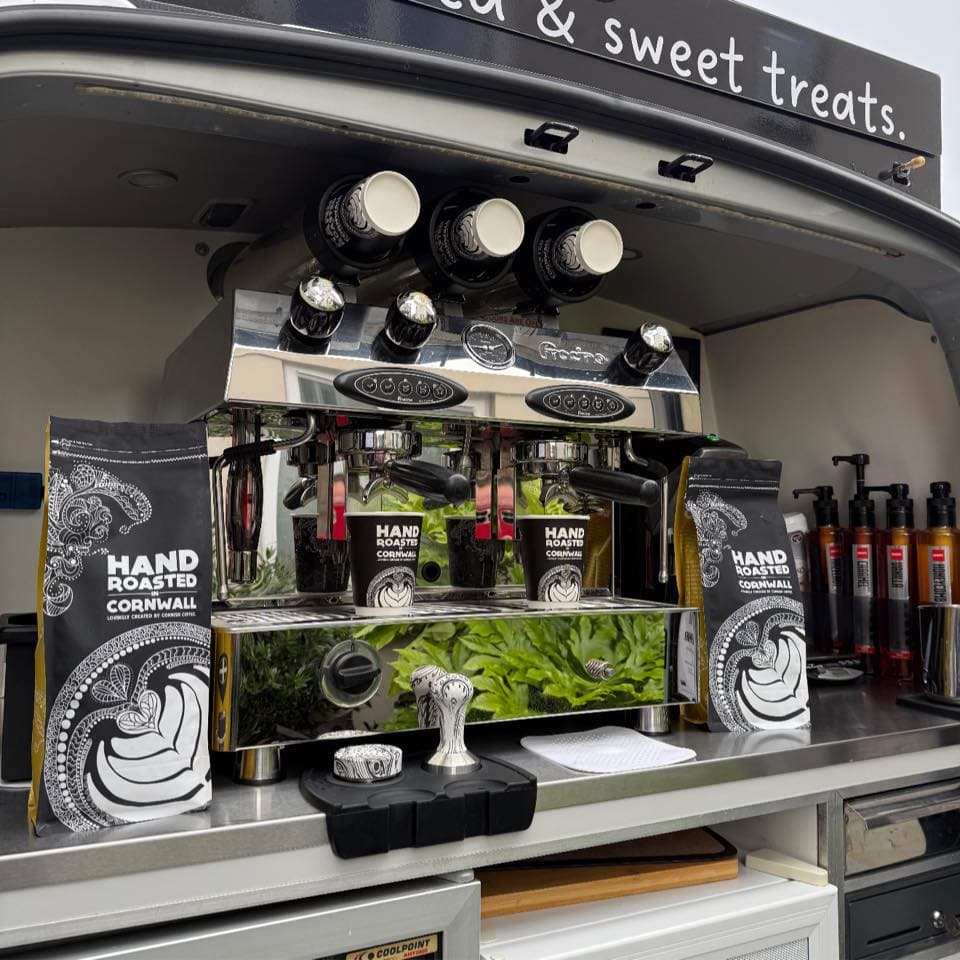 Two-group espresso machine inside The Roaming Barista van flanked by Cornish Coffee 'Hand Roasted in Cornwall' bags, with the 'tea & sweet treats.' canopy banner above