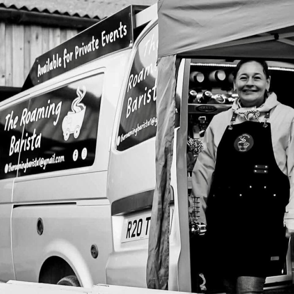 Shelley, owner of The Roaming Barista, smiling from inside the side-door coffee bar of her tuxedo-livery van